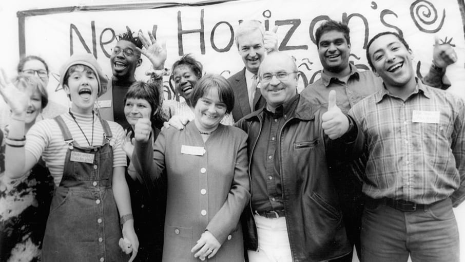Black and white photo of New Horizon group members smiling and with their thumbs up posing in front of a hand made New Horizon banner. Journalist Jon Show positioned centre back.